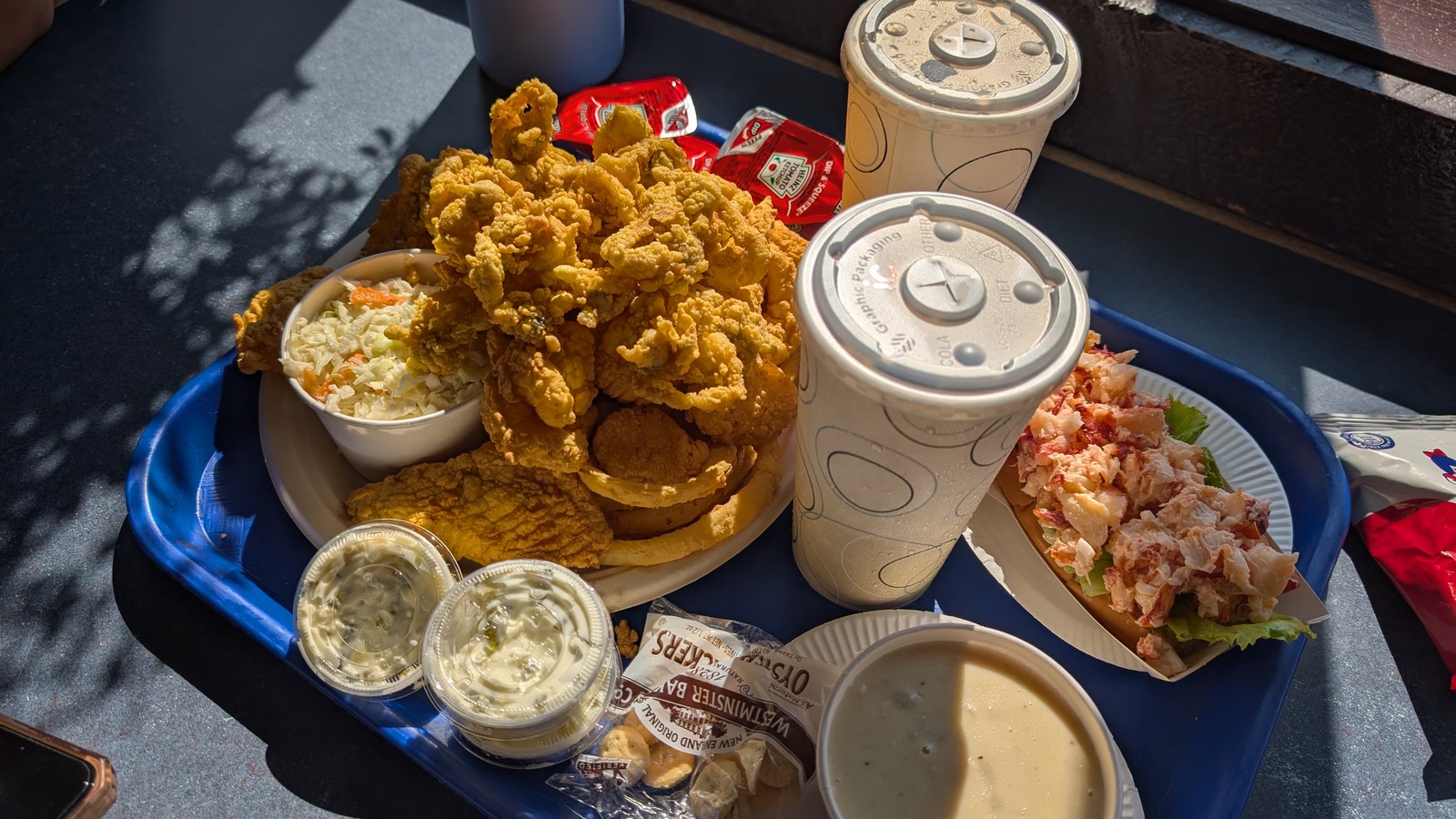 Blue tray loaded with fried seafood, chowder, tartar sauce, and a lobster roll by the window.