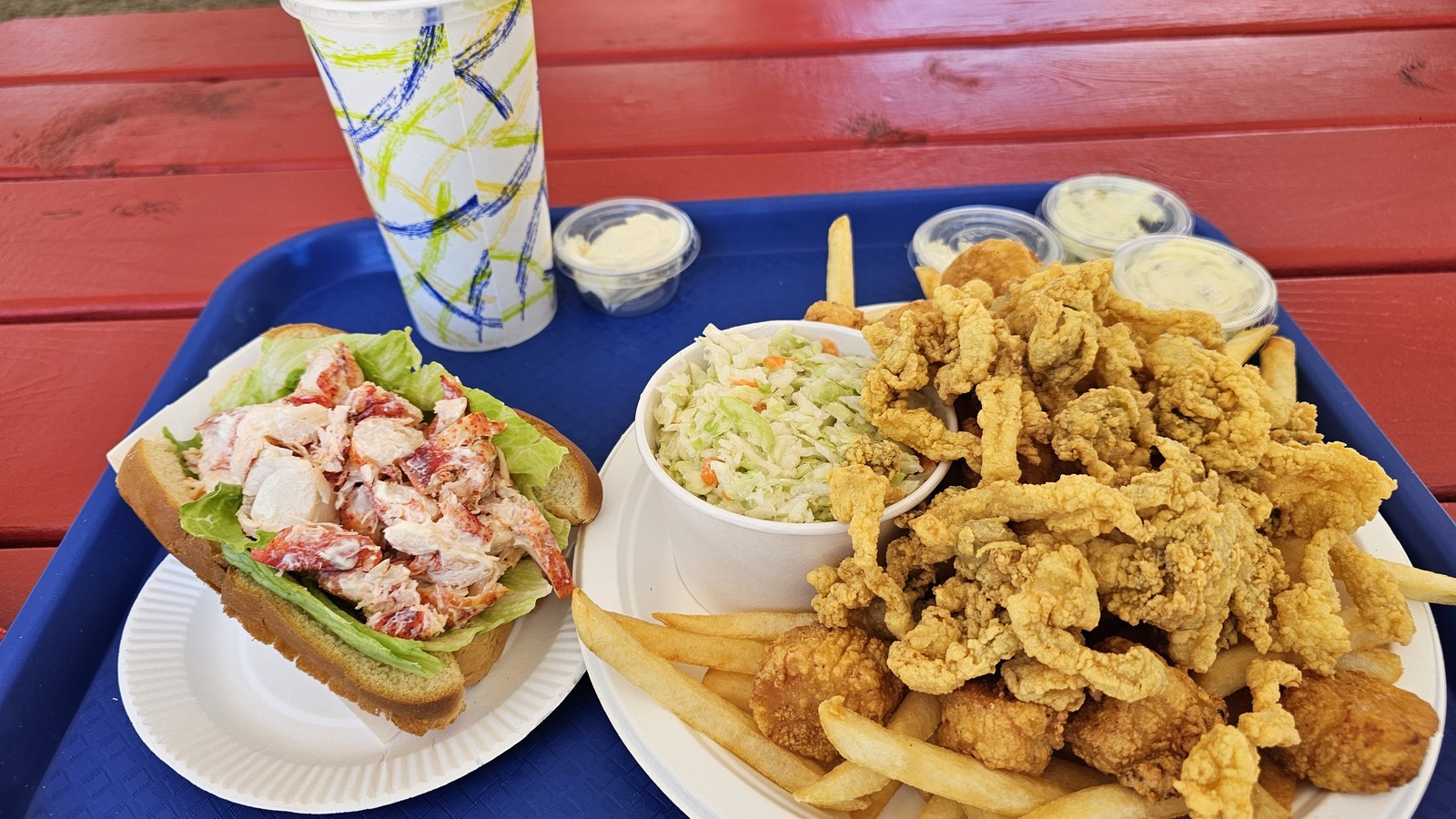 Lobster roll and fried clams platter on a blue tray set against a red picnic table.