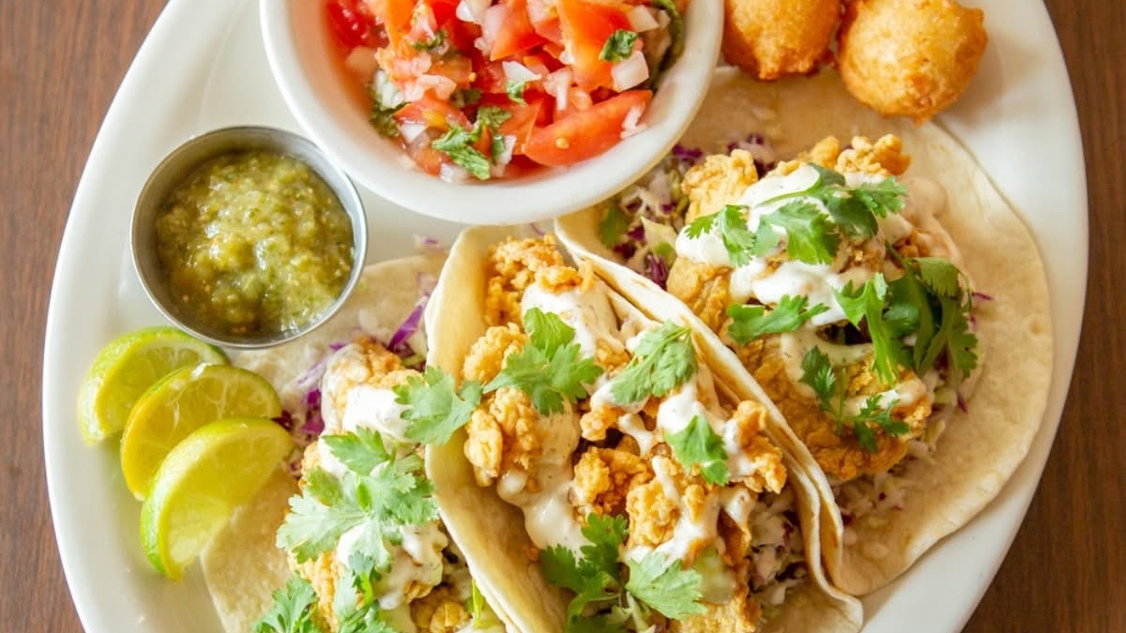 Three fish tacos with pico de gallo, salsa verde, lime wedges, and hush puppies