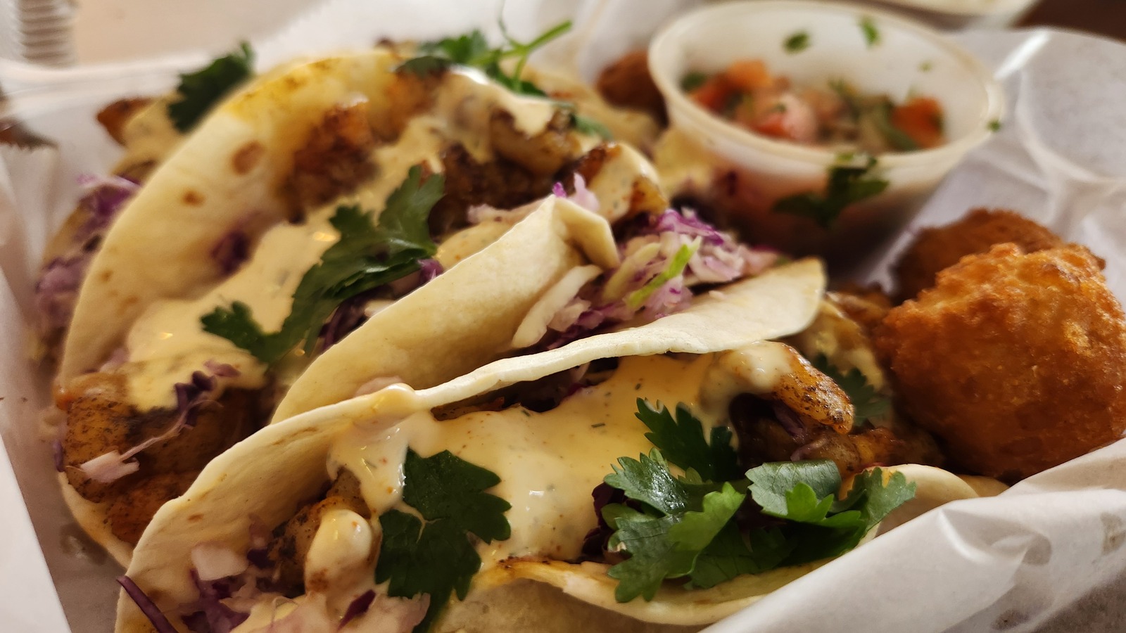 Close-up of fish tacos with creamy sauce, slaw, and hush puppies