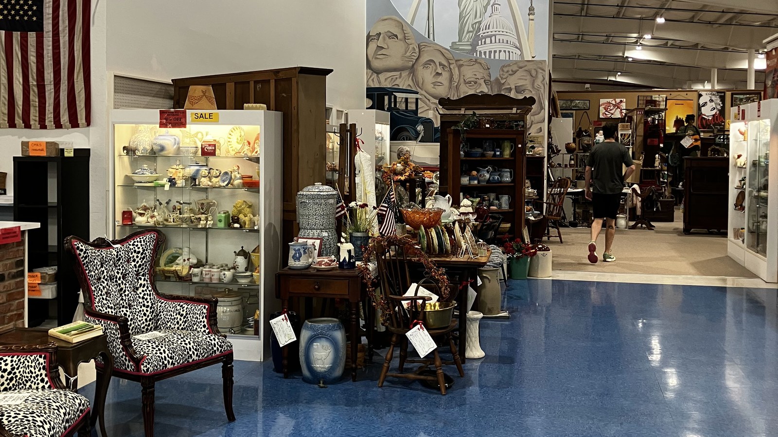 Blue-floored aisle at Exit 76 Antique Mall with patriotic flags, mural art, furniture, and lit display cases