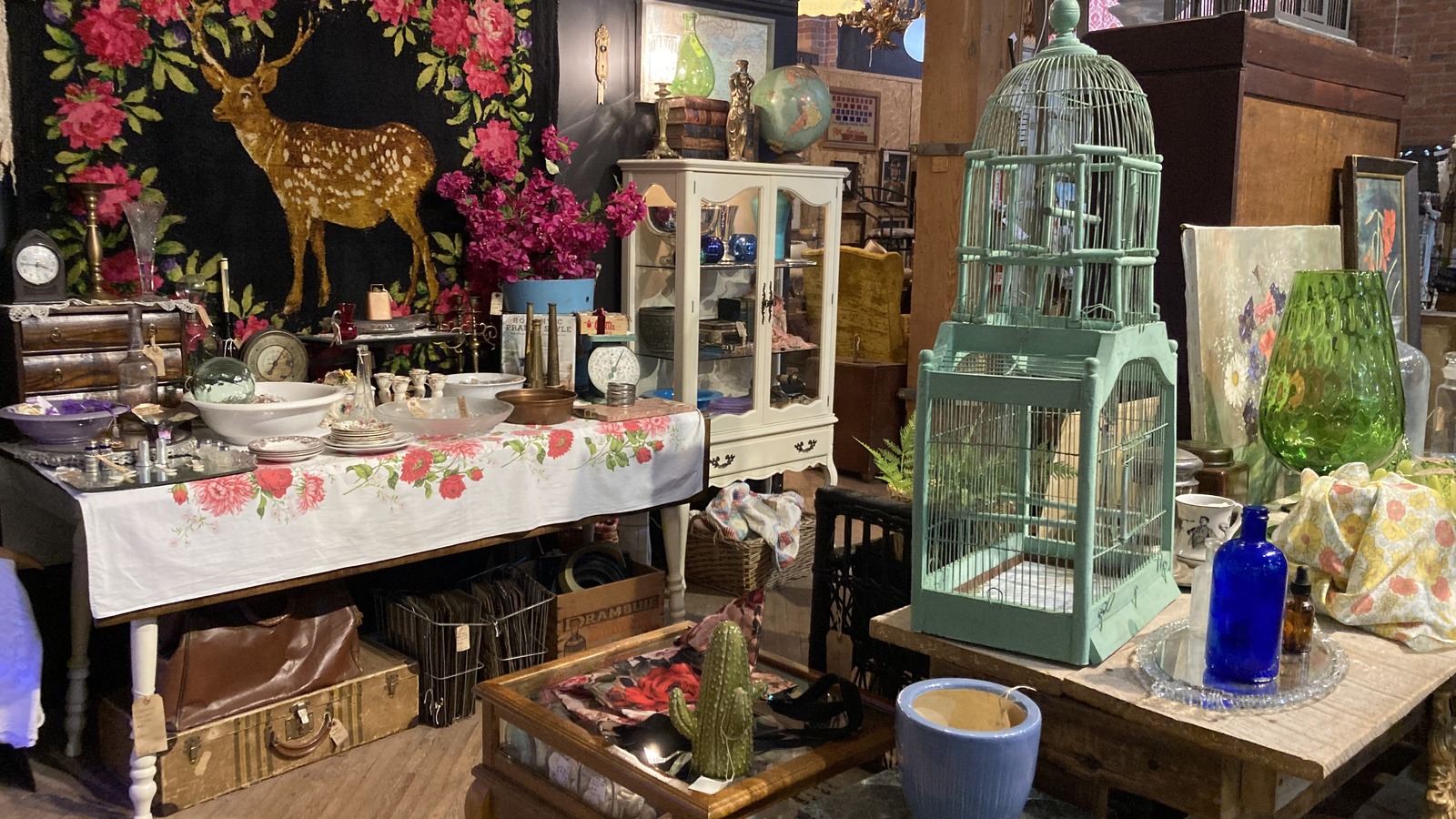 Colorful decorative booth with a floral deer textile, painted birdcage, and vintage furniture at Midland Arts & Antiques Market