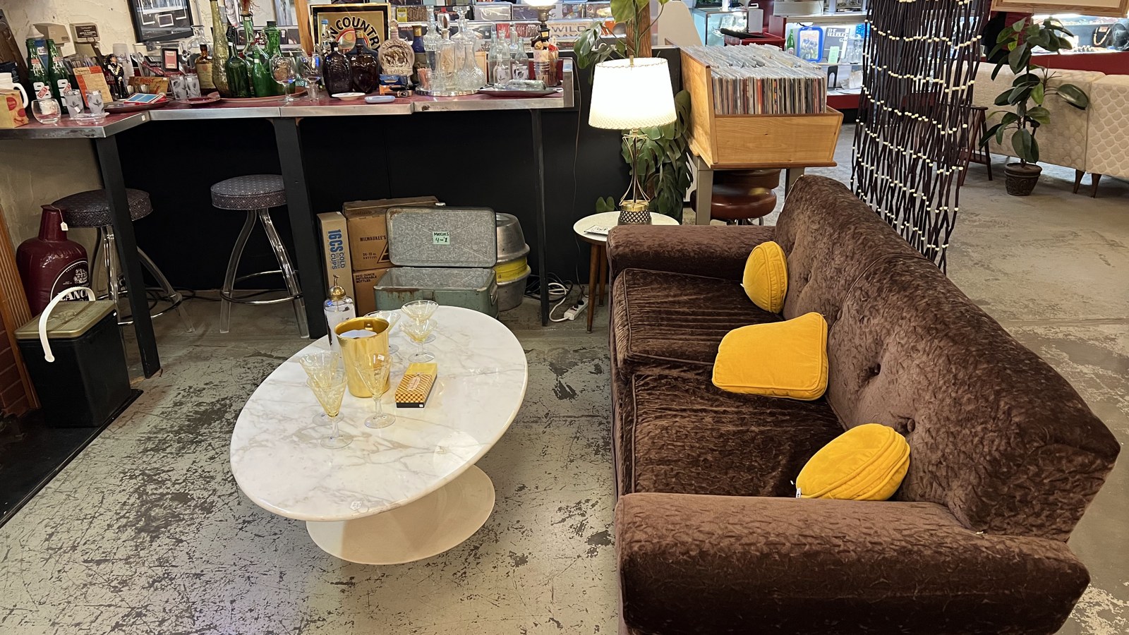 Retro lounge corner with a sofa, lamp, barware, and nearby record bins inside The Bomb Shelter