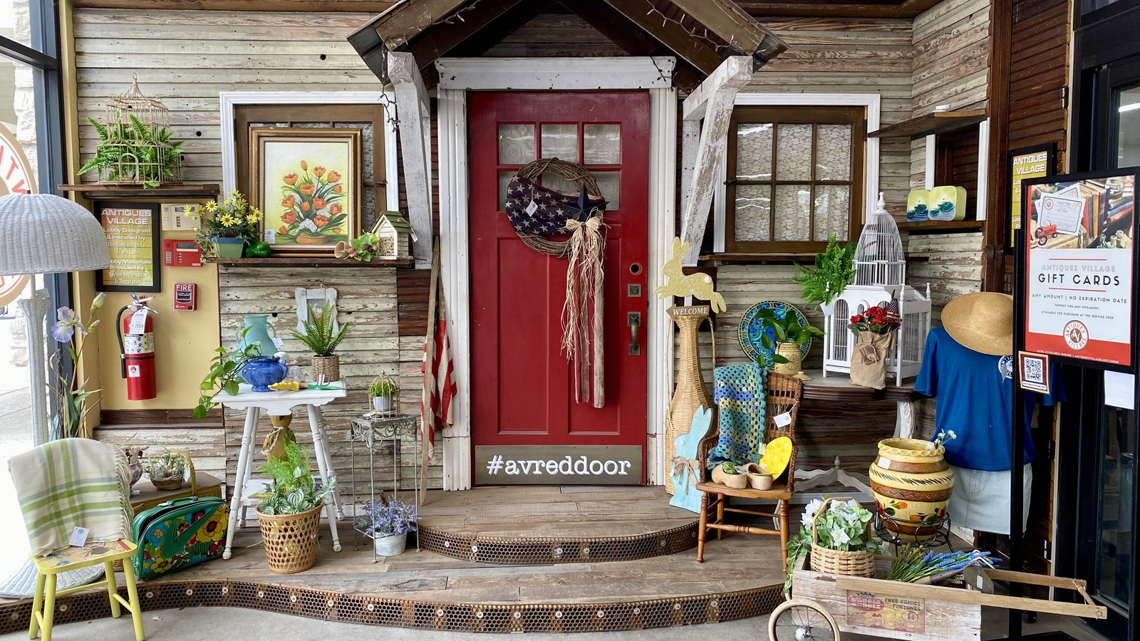 Rustic front display with a red door, plants, framed art, and vintage accents inside Antiques Village